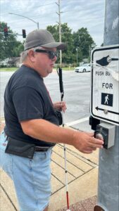 Person standing at a street corner and pressing a pedestrian crossing button while holding a long white cane, with traffic signals and an intersection visible nearby.