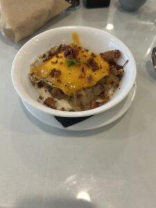 A bowl of grits topped with cheese, bacon, green onions, and potatoes sits on a white tablecloth