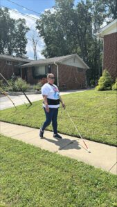 A person in a white tshirt walks down the sidewalk with their white cane extended in front of them. Behind them are brick houses and green lawns.