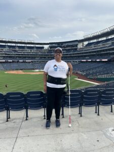 A person in a white tshirt and baseball cap smiles wide and poses with a white cane in hand. They are standing in the stands of a baseball stadium with the ballfield down below in the background.