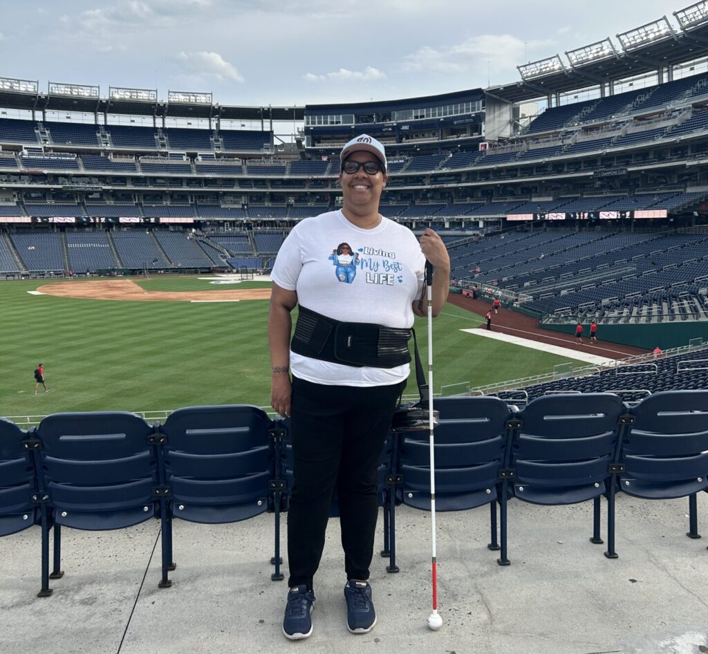 A person in a white tshirt and baseball cap smiles wide and poses with a white cane in hand. They are standing in the stands of a baseball stadium with the ballfield down below in the background.
