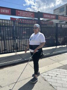 A person with a white cane walks outside the gate of a baseball stadium. They are wearing sunglasses and a backwards baseball cap. Through the gate the sign reads "Nationals Park".