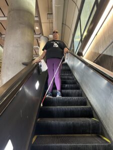 A person in a black shirt and purple pants stands on an escalator moving downwards. One hand in on the railing, and the other holds a white cane that rests just above the step below.