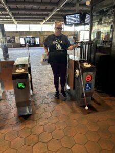 A person dressed in all black is exiting a train station, walking through turnstiles with their white cane extended in front of them