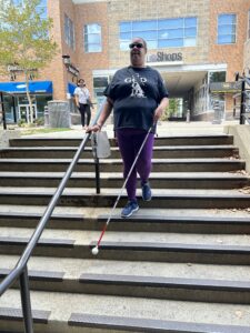 A person walks down a set of concrete stairs with their white cane extended on the stairs in front of them. They are outdoors in front of a row of shops.