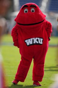 Red, fuzzy WKU Hilltopper mascot standing on a grassy sports field with hands on hips, featuring large cartoon eyes and the letters “WKU” displayed on the front of the costume.