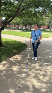Person using a white mobility cane walks along a campus sidewalk beneath a leafy tree, scanning the path ahead. Brick buildings, grass, and additional trees are visible in the background.