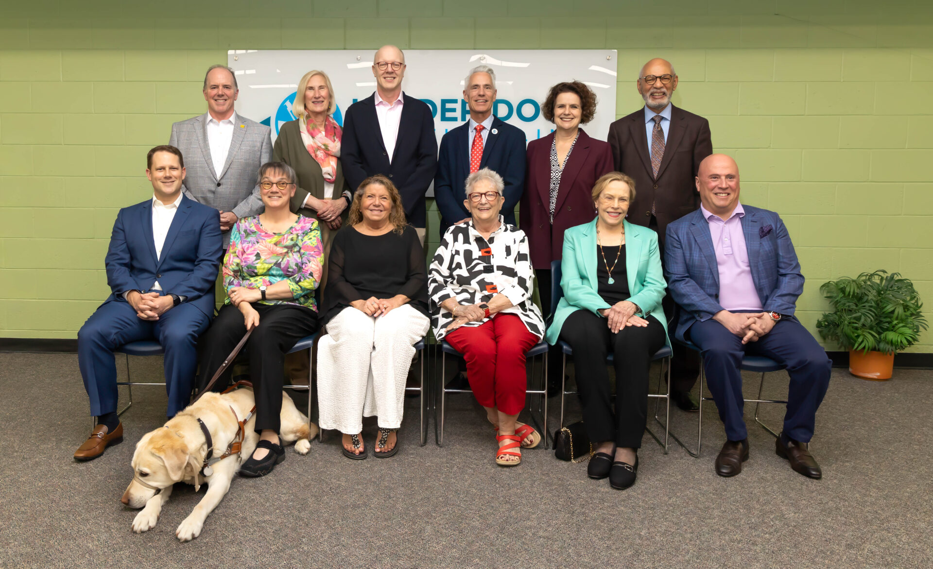 Group portrait of 12 people posed in two rows at an indoor event, with a yellow Labrador wearing a working harness lying in front