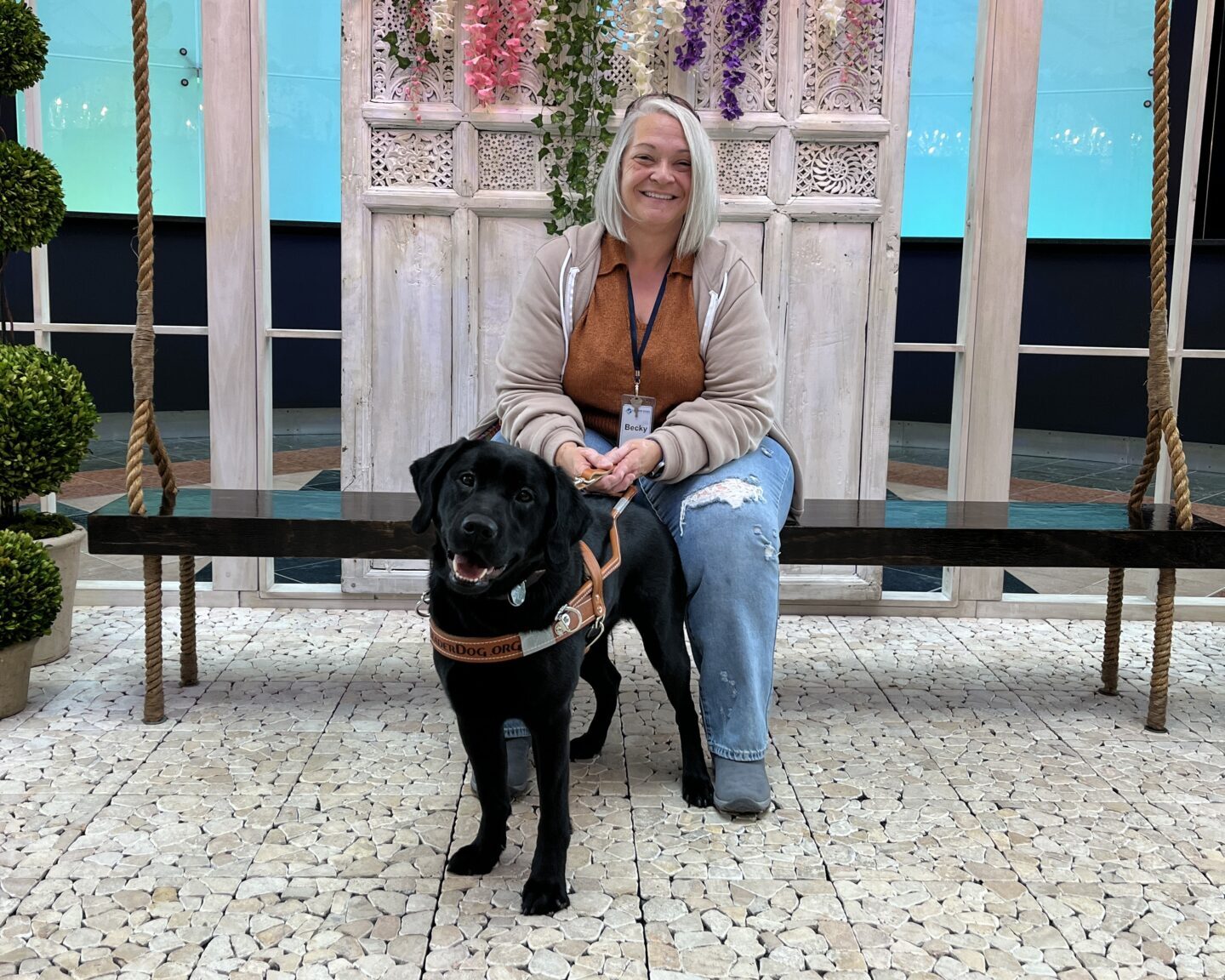 A person sits on a bench smiling while holding the leash of a black lab Leader Dog.