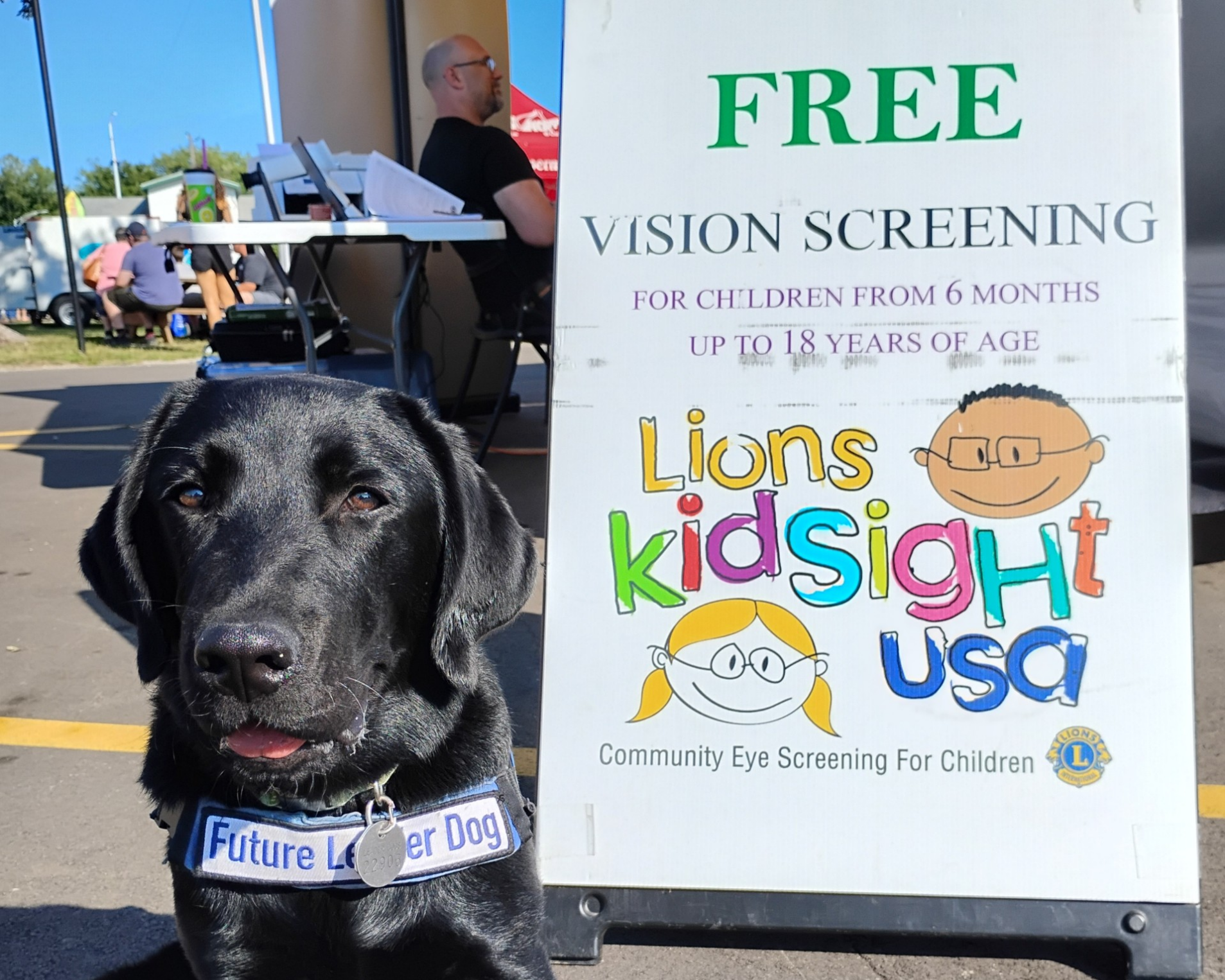 A black Labrador looks toward the camera with their togue out while wearing a Future Leader Dog vest. Behind the dog you see a sign for Free Vision Screenings for children from 6 months to up to 18 years. With the Lions Kidsight USA logo and the Lion emblem.