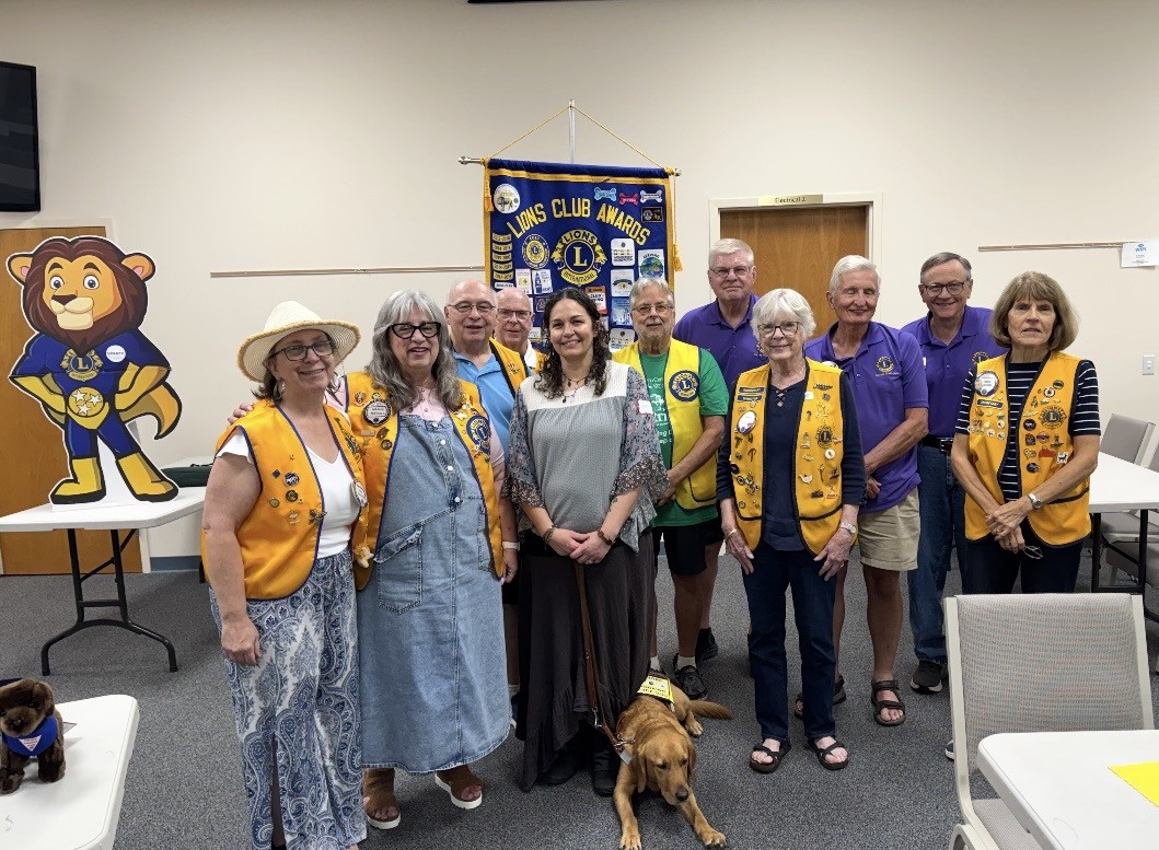 Group of Lions Club members and guests posing at a club gathering, with a Lions Club awards banner, lion mascot display, and dog.