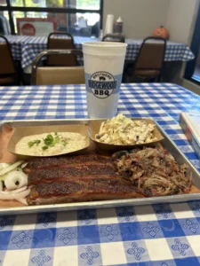 A butcher paper-lined silver tray on a blue gingham tablecloth holds barbecued ribs, pulled pork, grits, and cole slaw.