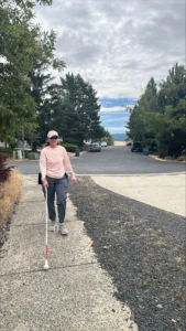 Wearing a pink top and dark pants, a person uses her white cane to walk up another hill. In the background are mountains. Beautiful, beautiful mountains.