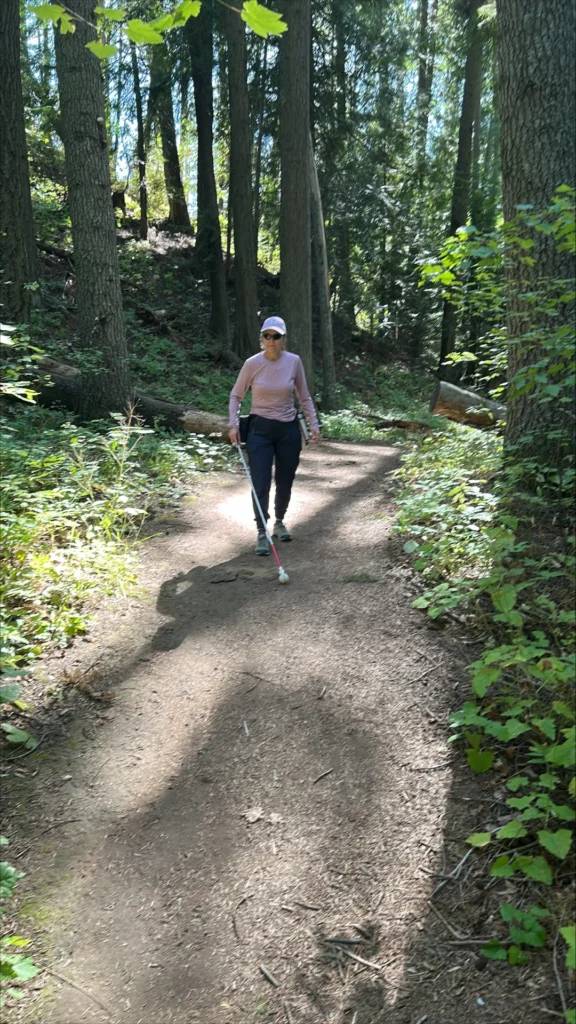 A person uses her white cane to navigate a dirt trail in a beautiful pine forest.