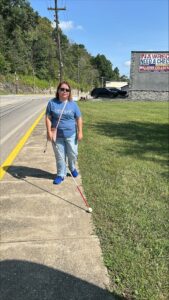 Wearing a light blue shirt and jeans, a person uses a white cane on the sidewalk in Olive Hill.