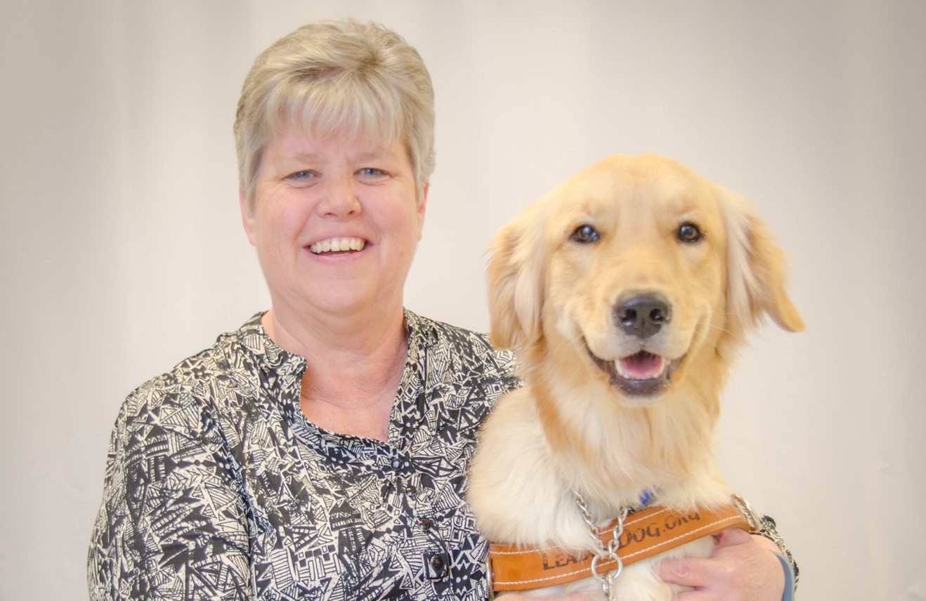 Photo of a woman sitting with her face toward the camera. She is wearing a flowery button up shirt and smiling. She has her arm around the golden retriever in Leader Dog harness sitting next to her.