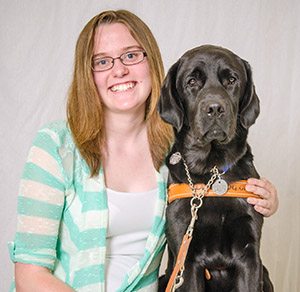 Photo of Shannon Columb, a young woman wearing glasses, a white shirt and a green and white-striped cardigan. She is seated and facing the camera, smiling. Her left arm is around a large black lab sitting on her left. He is wearing his leather Leader Dog harness.