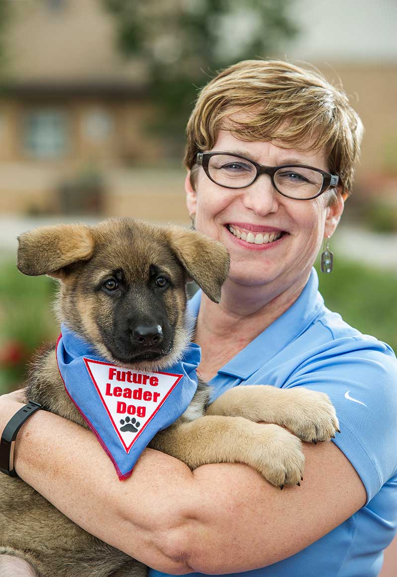 Closeup of Lorene Suidan smiling and holding a young German shepherd puppy wearing Future Leader Dog bandanna
