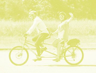 Green colored image of a man and a teenage girl on a tandem bike. Both of them are smiling and the girl uses one hand to wave toward the camera