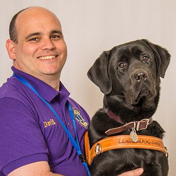 Photo of a man in a purple polo shirt with the Lions Clubs logo on it. He is smiling at the camera and sitting in front of a gray backdrop with his hands on the black lab in harness seated next to him