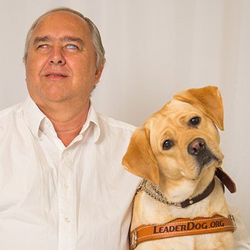 Photo of a man in a white button up shirt sitting in front of a gray backdrop. Next to him is a yellow lab in leather Leader Dog harness with her head tilted toward Francisco