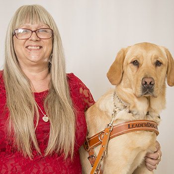 Photo of a woman wearing a red dress smiling and facing the camera. She is sitting in front of a gray backdrop with her left arm around a yellow lab/golden retriever cross in leather Leader Dog harness
