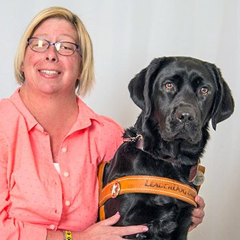 Photo of a woman in a pink button up shirt sitting in front of a gray backdrop and smiling at the camera. Her arms are around the black lab seated to her left
