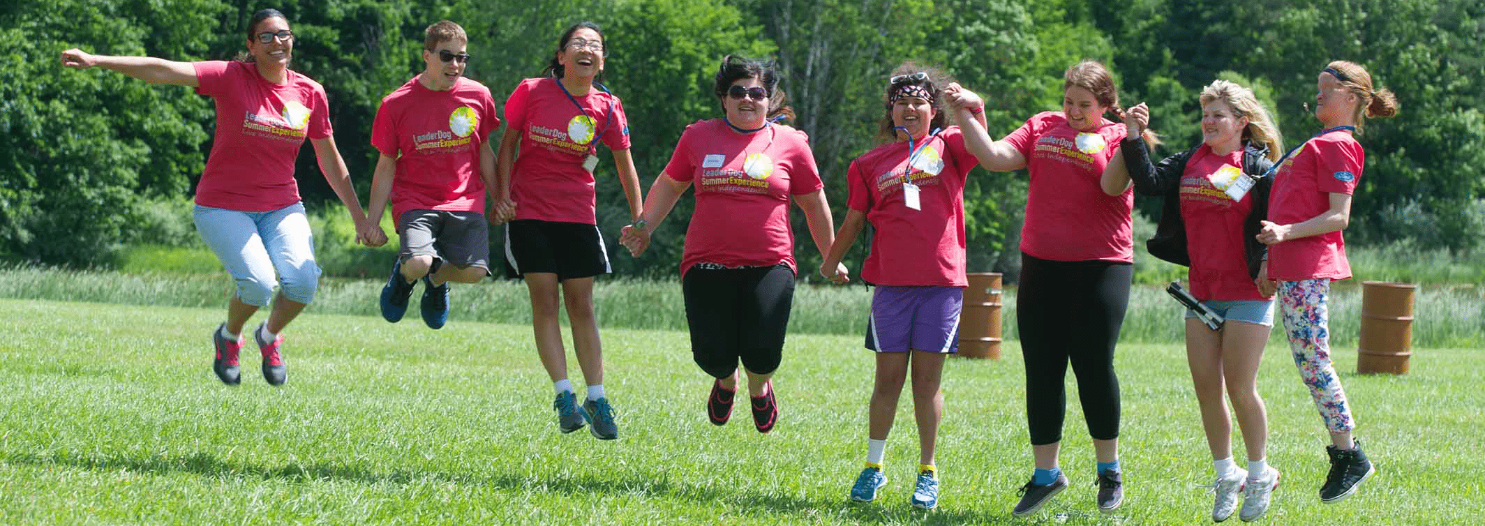 8 teenagers stand in a line holding hands in the middle of a field with trees in the background. Each wears a red summer experience camp tee shirt. The image captures them mid jump as they smile facing the camera.