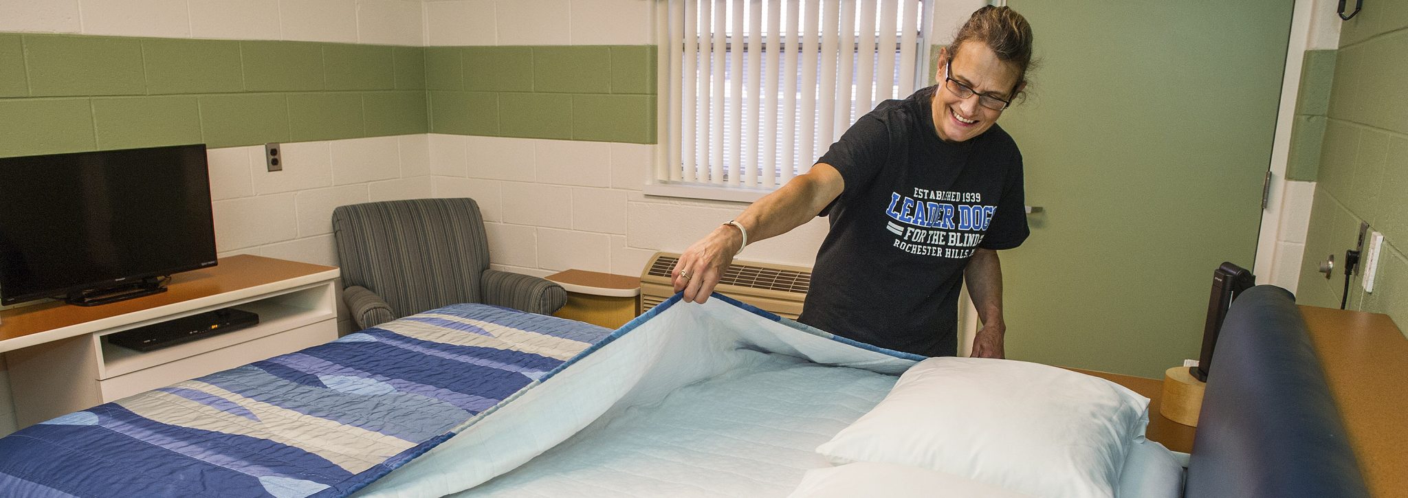 A woman smiles while pulling a blanket over a neatly made bed