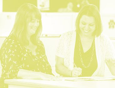 Green colored image of two women standing behind a table or desk. They are smiling and looking down at some papers while one of them holds a pen