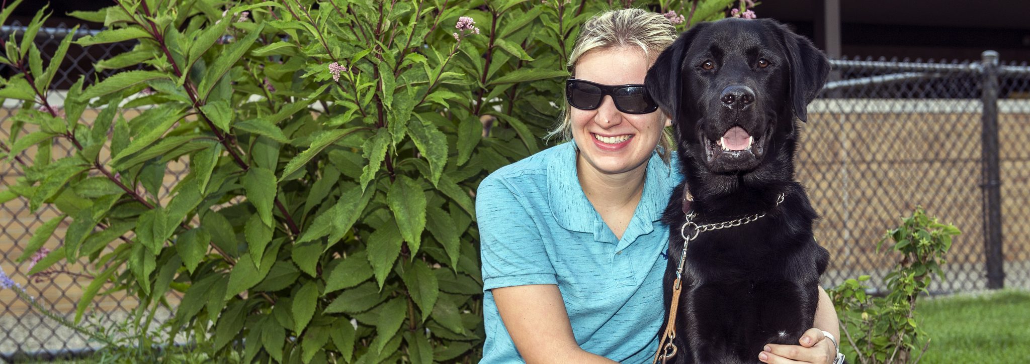 Photo of a smiling woman wearing sunglasses and sitting or kneeling in front of a bush. She has her arms around the black Labrador sitting next to her. The dog is sitting and looking directly at the camera with tongue slightly out.
