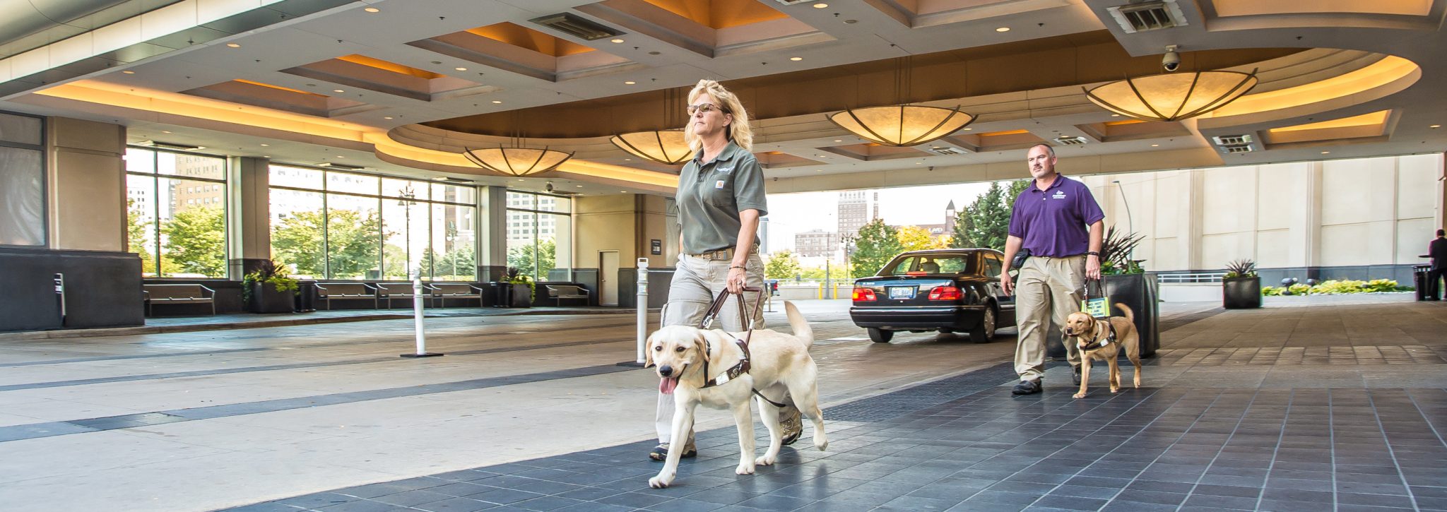 A woman walks with a guide dog in harness across a covered parking area in front of the MGM Grand Detroit. Behind her, a man walks with another dog in harness