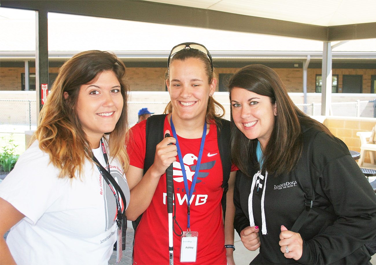 Three young women face the camera, smiling. The woman in the center is holding a white cane