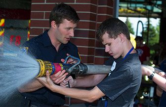 A fireman assists teenager Brock with holding a fire hose that is spraying water