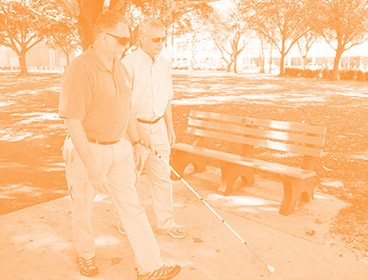 Orange colored image of two men walking down a sidewalk with a park bench and trees in the background. One man is holding a white cane
