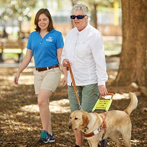 Two women, one older and one younger, walk outdoors on a sunny day. The older woman is walking with a yellow lab guide dog in harness. The younger woman is beside them, smiling and observing