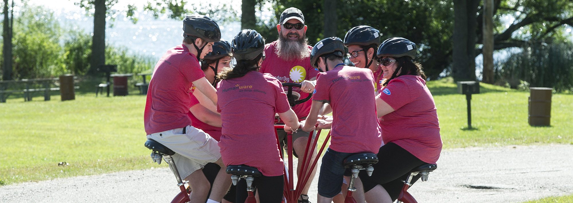 Six teens wearing helmets and an adult man sit in a circle on a group bike outside on a sunny day. They are smiling and pedaling