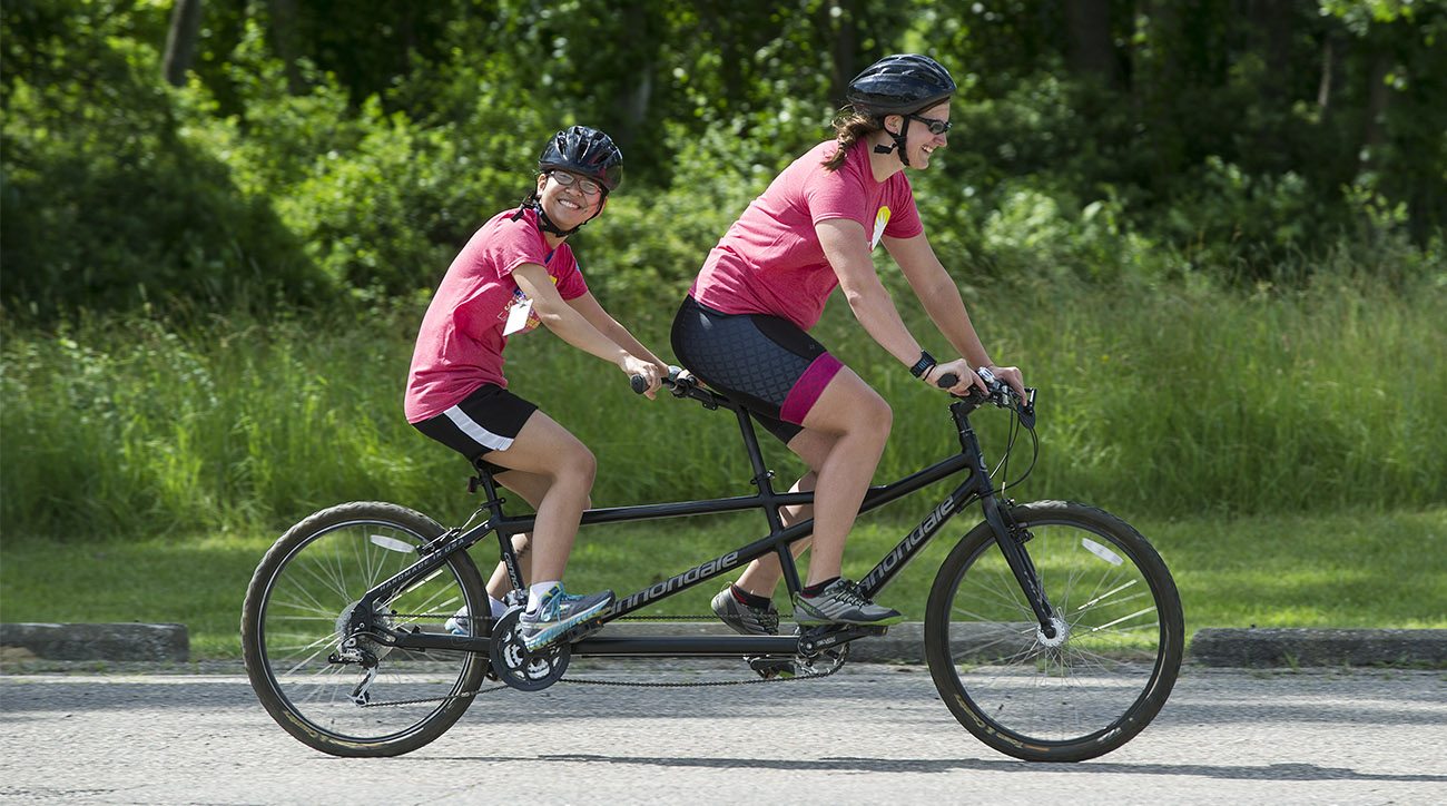 An adult woman, sitting in front, and a teenage girl ride a tandem bike on a sunny day on a road in front of trees and bushes. Both are smiling