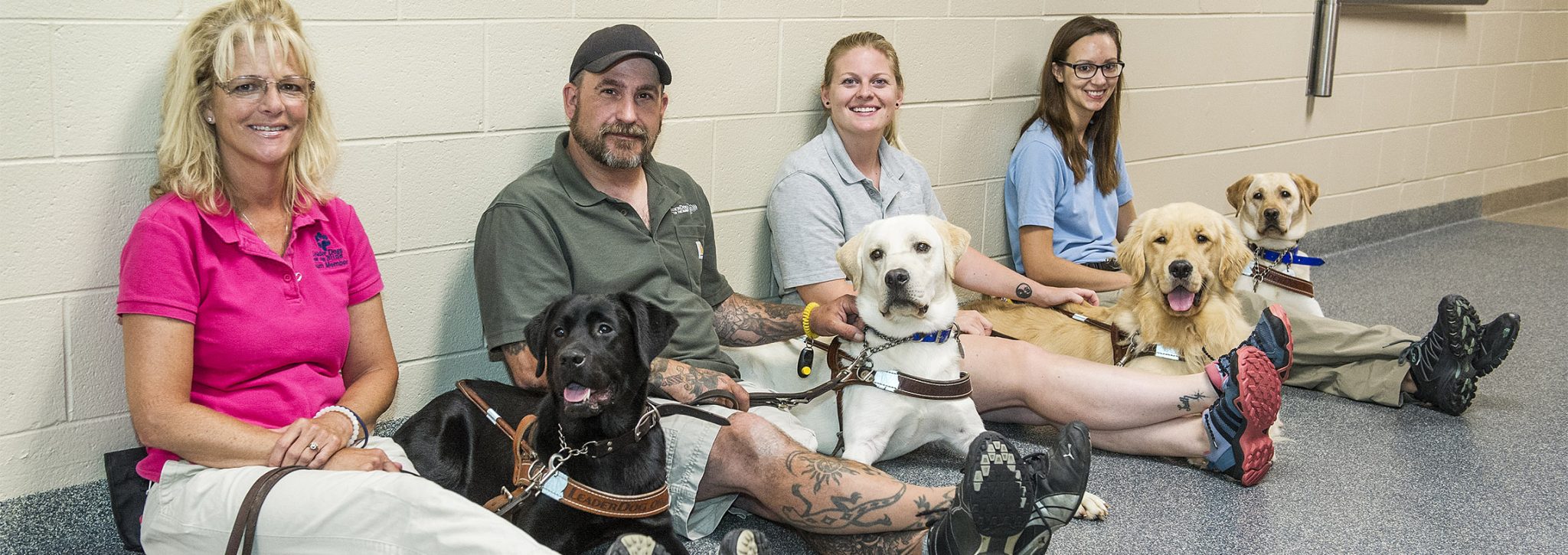 Four Leader Dog instructors, three women and one man, sit with legs outstretched on the floor in a hallway. Beside each one of them is a lab or golden retriever in harness