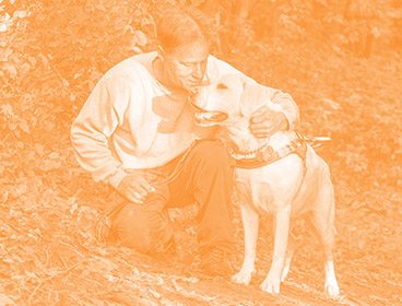 Orange colored image of a man kneeling on a dirt path with his arm around a yellow lab in Leader Dog harness