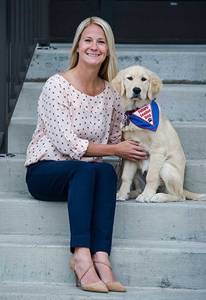 Dani Landolt sits on concrete steps with her hands on an adolescent golden retriever puppy wearing Future Leader Dog bandanna