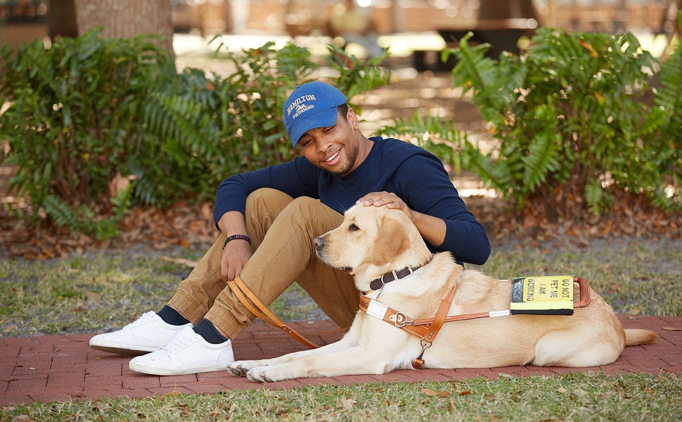 Gui sits on a brick path outdoors on a sunny day. In one hand he holds the leash of the yellow lab and golden retriever cross lying beside him in harness, Emma. With his other hand, he pets Emma