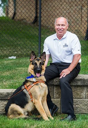 Rod Haneline sits outdoors on a low wall smiling and holding the leash of a seated German shepherd in harness