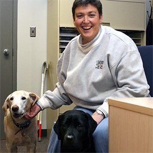A smiling Carrie sits at a desk. She is scratching the chin of a yellow lab standing next to her while a black lab sits in front of her legs