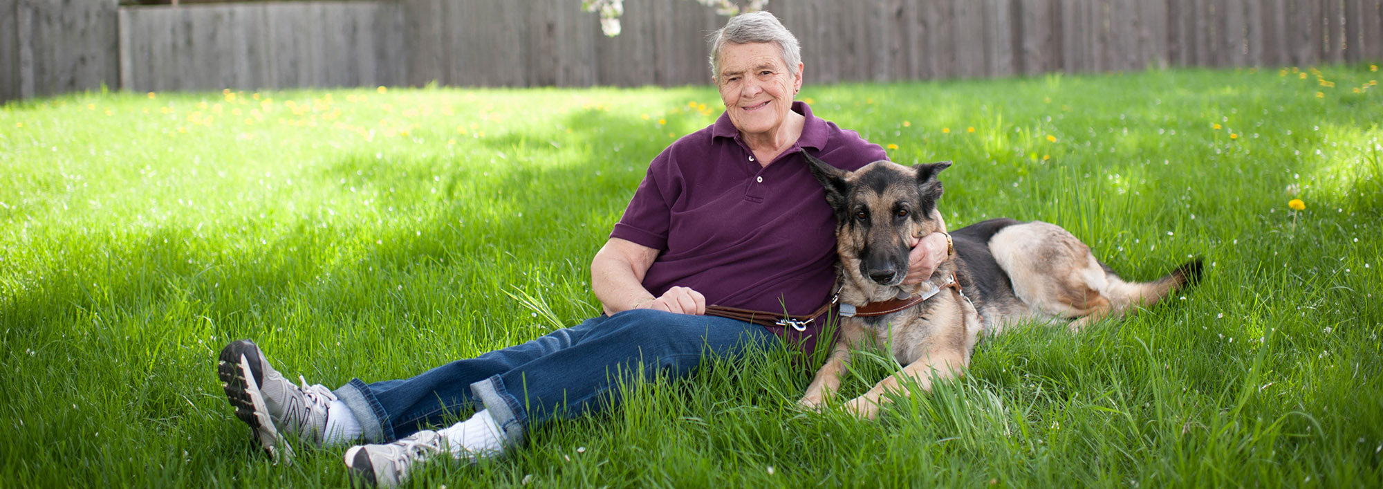 An older woman sits on grass on a sunny day, smiling at the camera. Her arm is around a German shepherd lying next to her. A wooden fence is visible in the background