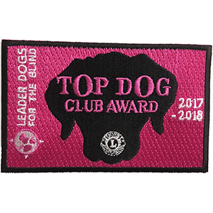 A pink rectangular patch with a black border. In the center of the patch is a black silhouette of a dog head and the Lions International logo. The patch features embroidered text, "Leader Dogs for the Blind, Top Dog Club Award 2017-2018"