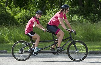 An adult woman, sitting in front, and a teenage girl ride a tandem bike on a sunny day on a road in front of trees and bushes. Both are smiling
