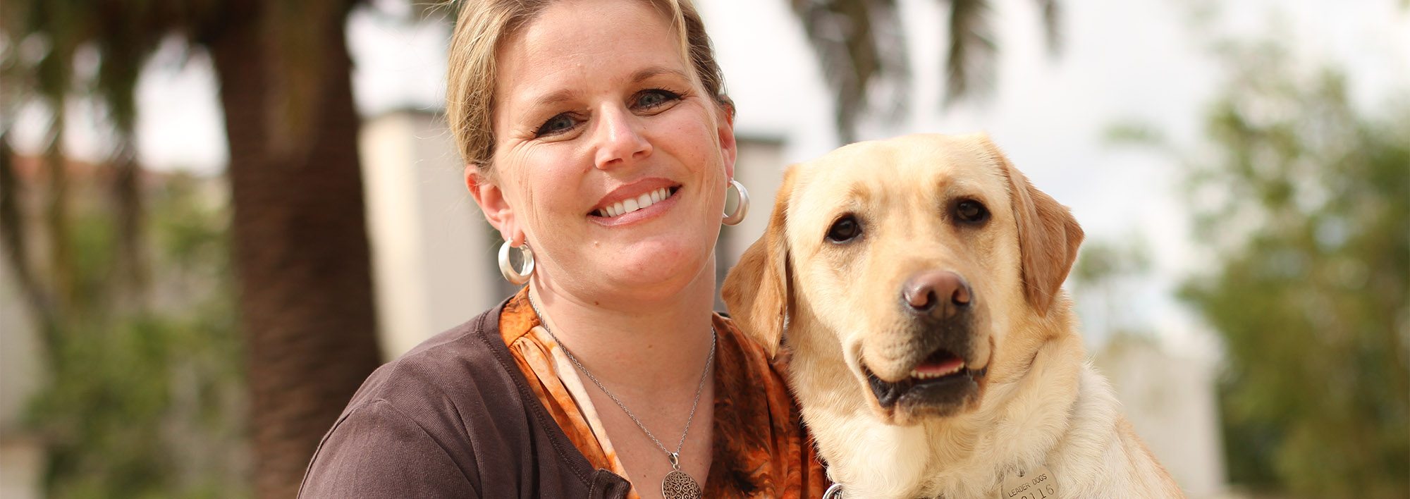 A woman sits outside on a sunny day smiling at the camera. Next to her is a yellow lab also looking into the camera