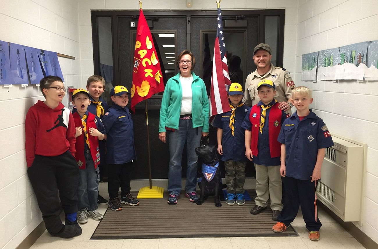 Mary stands holding a black labrador's leash in a hallway in front of double doors. She is smiling at the camera and surrounded on both sides by young boys wearing Boy Scouts shirts and hats. A Scout leader is also standing with them