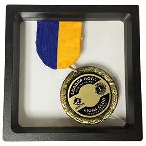 A medallion sits in a box with a yellow and blue ribbon attached,. The medallion is inscribed with a Leader Dogs for the Blind logo and Lions Club International logo.